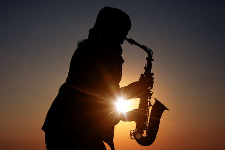 Silhouette of woman playing saxophone against beautiful sky at sunsetの写真素材