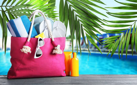 Bag with different beach accessories on gray wooden surface and green palm leaves near outdoor...