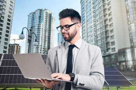 Young businessman with laptop near solar panels and beautiful view of cityscape. alternative energy sourcesの写真素材