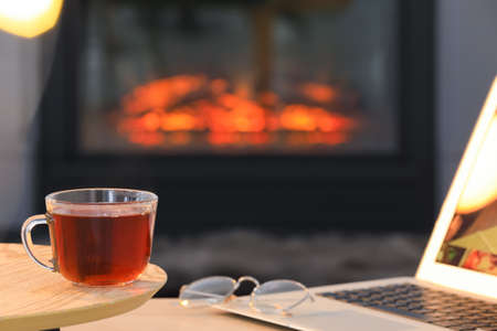 Glass cup of hot tea on wooden table near fireplace at home, space for text. Cozy atmosphereの写真素材
