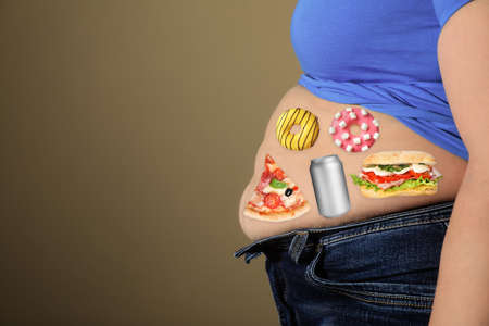 Overweight woman in tight shirt and jeans with images of different unhealthy food on her belly against light brown background, closeup. Space for textの写真素材