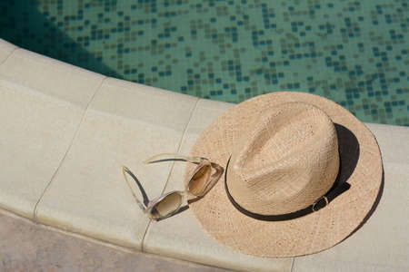 Stylish hat and sunglasses near outdoor swimming pool on sunny day, space for text. beach accessoriesの写真素材