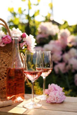 Bottle and glasses of rose wine near beautiful peonies on wooden table in gardenの写真素材