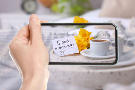 Woman taking picture of aromatic coffee, flowers and card with phrase GOOD MORNING! on tray, close...