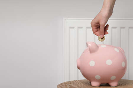 Woman putting coin into piggy bank near heating radiator, closeup. Space for textの写真素材