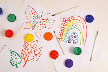 Cute children's drawings, brushes and set of paints on white table, flat lay