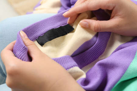 Woman holding colorful sweater with blank clothing label on blurred background, closeup