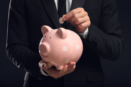 Businessman putting coin in piggy bank on black background, closeupの写真素材