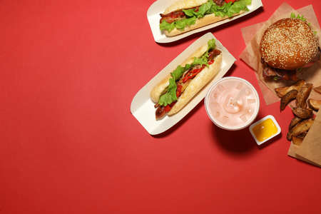 Tasty burger, hot dogs, potato wedges, sauce and refreshing drink on red background, flat lay with...
