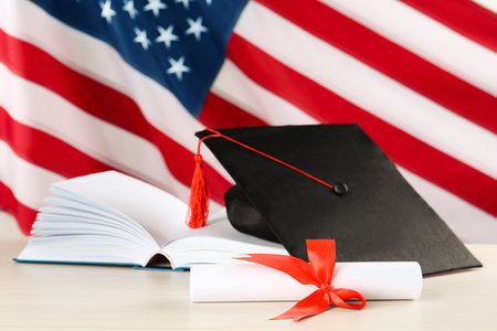 Black graduation hat, diploma and book on white wooden table against American flagの写真素材