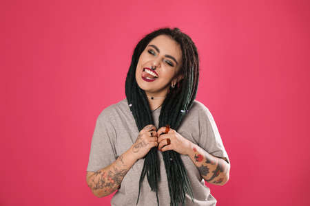 Beautiful young woman with tattoos on arms, nose piercing and dreadlocks against pink background