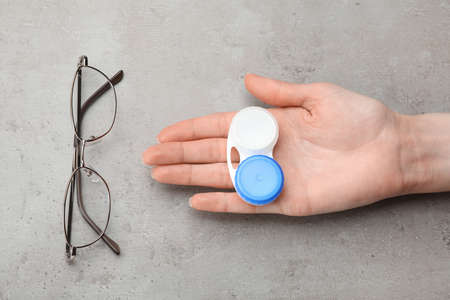 Woman holding case with contact lenses near glasses at gray table, top view