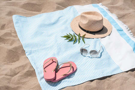 Beach towel with straw hat, sunglasses, leaves and flip flops on sandの写真素材