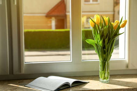 Bouquet of beautiful yellow tulip flowers in glass vase and open book on windowsill. Space for textの写真素材