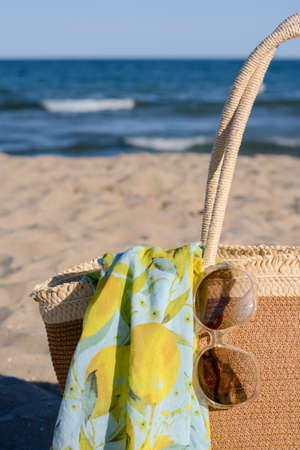 Straw bag with beach wrap and sunglasses on sandy seashore, closeup. summer accessoriesの写真素材