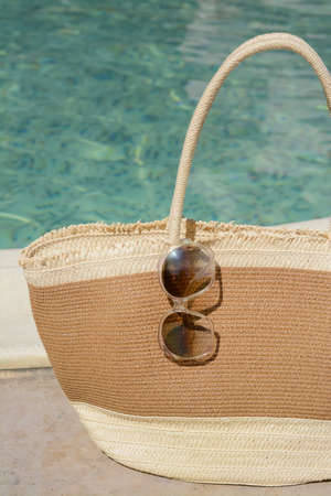 Stylish bag and sunglasses near outdoor swimming pool on sunny day. beach accessoriesの写真素材