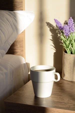 Cup of coffee on wooden night stand near bed in the morningの写真素材