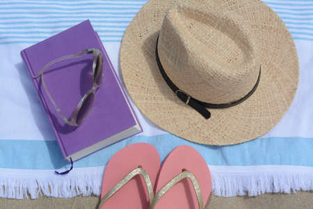 Beach towel with book, straw hat, sunglasses and flip flops on sand, flat layの写真素材