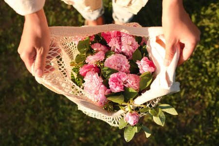 Woman holding mesh bag with beautiful tea roses outdoors, top view