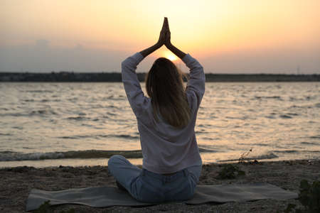 Young woman meditating on beach near river at sunset, back view