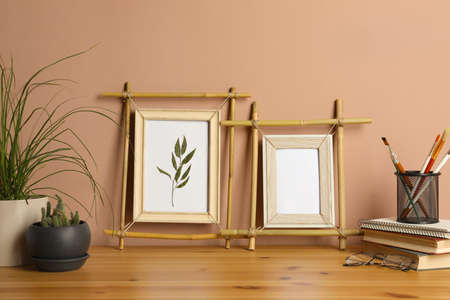 Stylish bamboo frames, potted houseplants and stationery on wooden table near brown wall