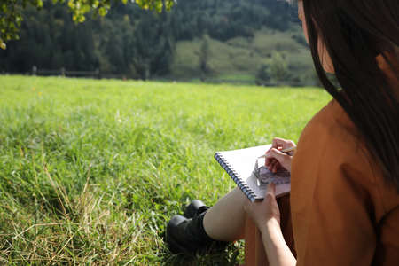 Beautiful young woman drawing with pencil in notepad outdoors on green grass, closeupの写真素材