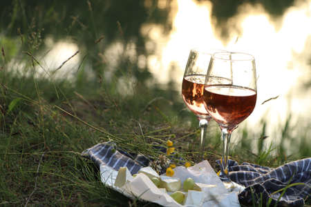 Glasses of delicious rose wine, cheese and grapes on picnic blanket near lake, space for textの写真素材