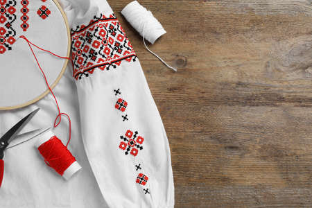 Shirt with red embroidery design in hoop, scissors and threads on wooden table, flat lay. National...