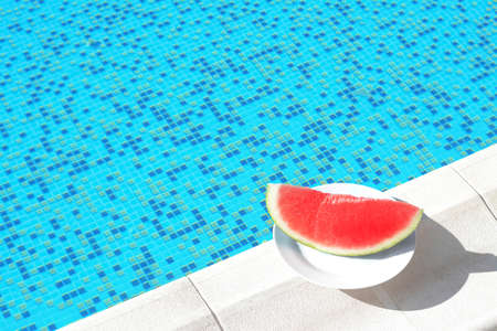 Slice of fresh juicy watermelon on white plate near swimming pool outdoors. Space for text