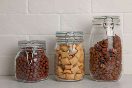 Glass containers with different breakfast cereals on white countertop near brick wall