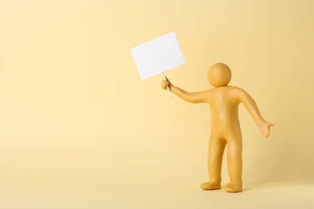 Human figure made of yellow plastic holding blank sign on beige background. Space for textの写真素材