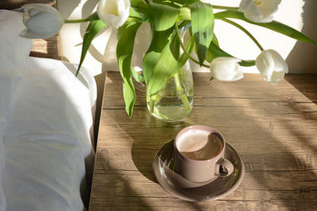 Beautiful white tulip bouquet and cup of coffee on nightstand in bedroomの写真素材