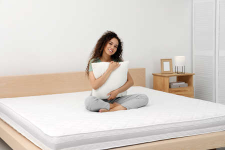 Happy young African American woman hugging pillow on bed with comfortable mattress at homeの写真素材