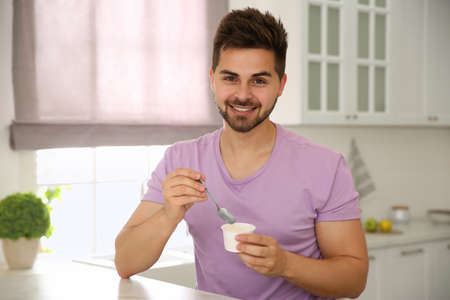 Happy young man with tasty yogurt at table in kitchenの写真素材