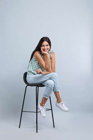 Beautiful young woman sitting on stool against light gray backgroundの写真素材