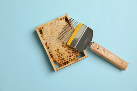 Hive frame with honeycomb and uncapping fork on light blue background, flat lay. Beekeepingの写真素材