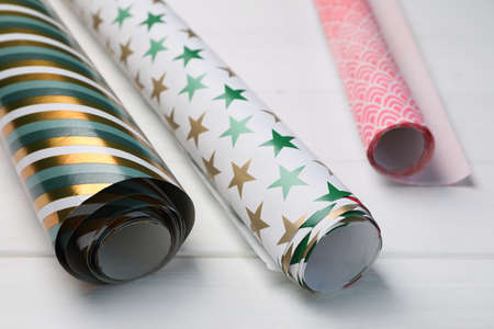 Different colorful wrapping paper rolls on white wooden table, closeup