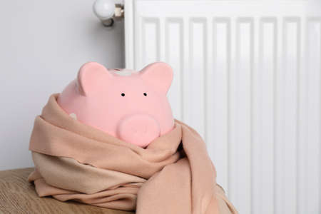 Piggy bank wrapped in scarf on wooden table near heating radiator. Space for textの写真素材