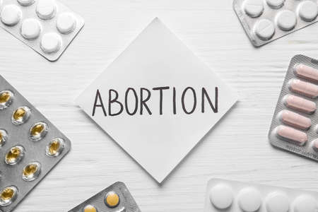 Paper note with word Abortion and pills on white wooden background, flat layの写真素材