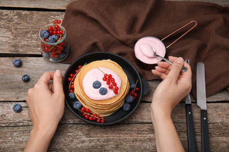 Woman putting natural yogurt onto tasty pancakes with blueberries and red currants at wooden table,...