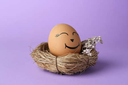 Egg with drawn happy face in nest on lilac background, closeupの写真素材
