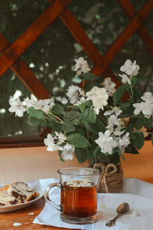 Glass cup of aromatic tea, tasty dessert and beautiful jasmine flowers on wooden table indoorsの写真素材
