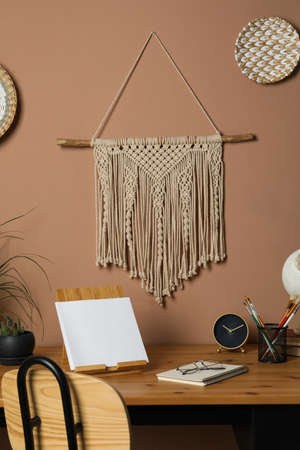 Stylish macrame and wicker wall decor hanging above wooden table with stationery