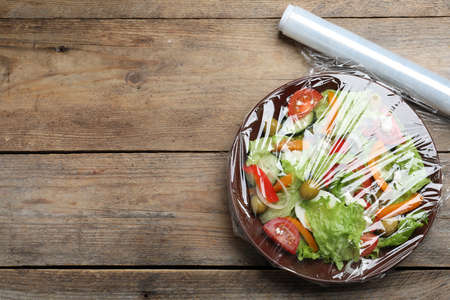 Bowl of fresh salad with plastic food wrap on wooden table, flat lay. Space for textの写真素材
