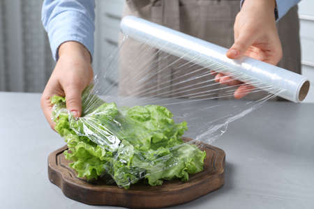 Woman putting plastic food wrap over fresh lettuce at light gray table indoors, closeupの写真素材