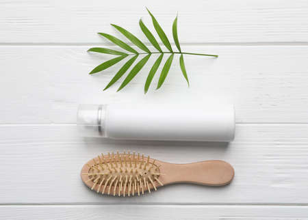 Dry shampoo spray, hairbrush and green twig on white wooden table, flat lay