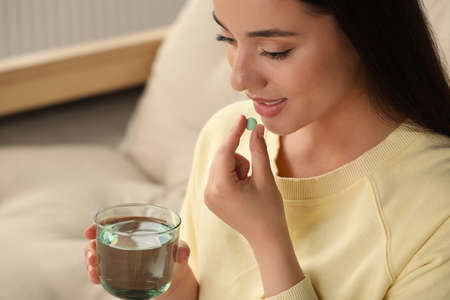 Young woman with glass of water taking dietary supplement pill indoors, space for text