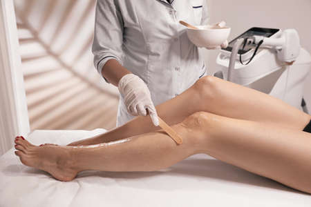 Professional cosmetologist applying gel on client's leg before laser epilation procedure in salon,...