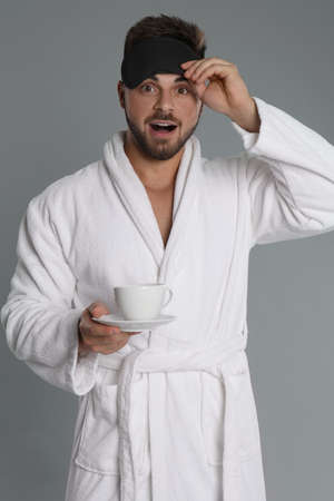 Young man in bathrobe with cup of coffee on gray backgroundの写真素材