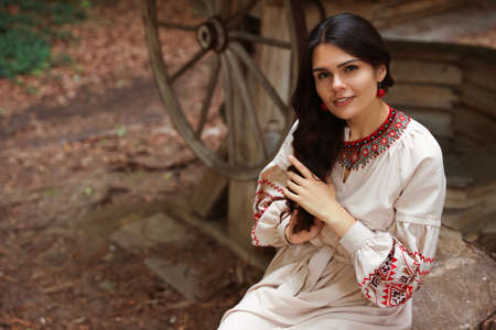 Beautiful woman wearing embroidered dress sitting near old wooden well in countryside. Ukrainian...